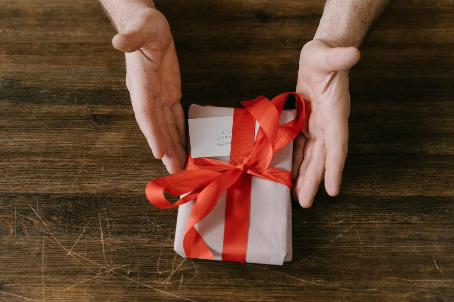 Hands cradle a small white gift box tied with a bright red ribbon on a wooden table surface.